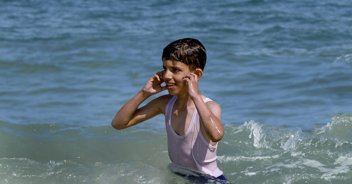 A boy in the sea cups his hands to his ears and smiles.